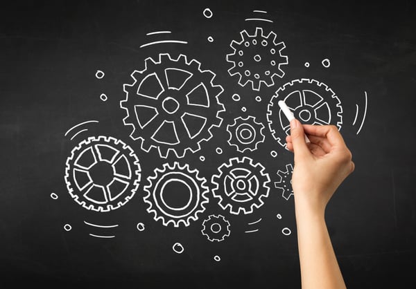 Female hand holding white chalk in front of a blackboard with gears drawn on it